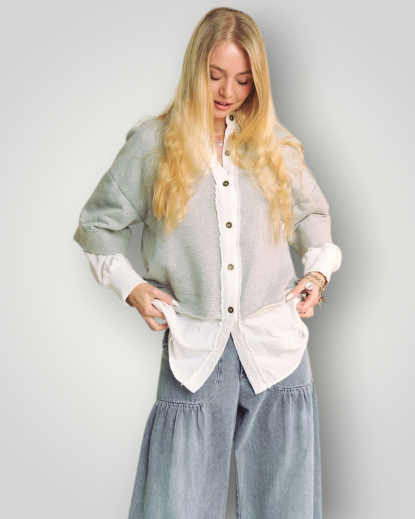 Woman wearing a layered outfit with a light gray cardigan, white shirt, and blue jeans against a plain background.