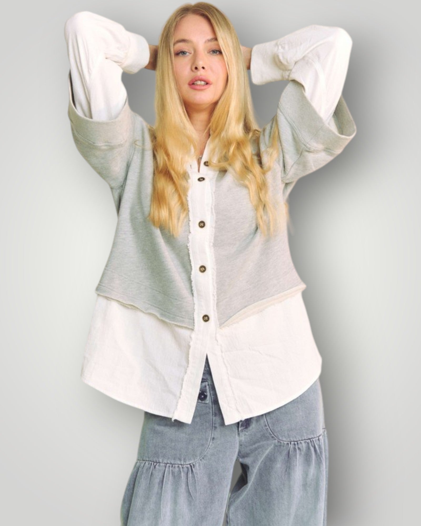 Woman wearing a layered outfit with a light gray cardigan, white shirt, and blue jeans against a plain background.