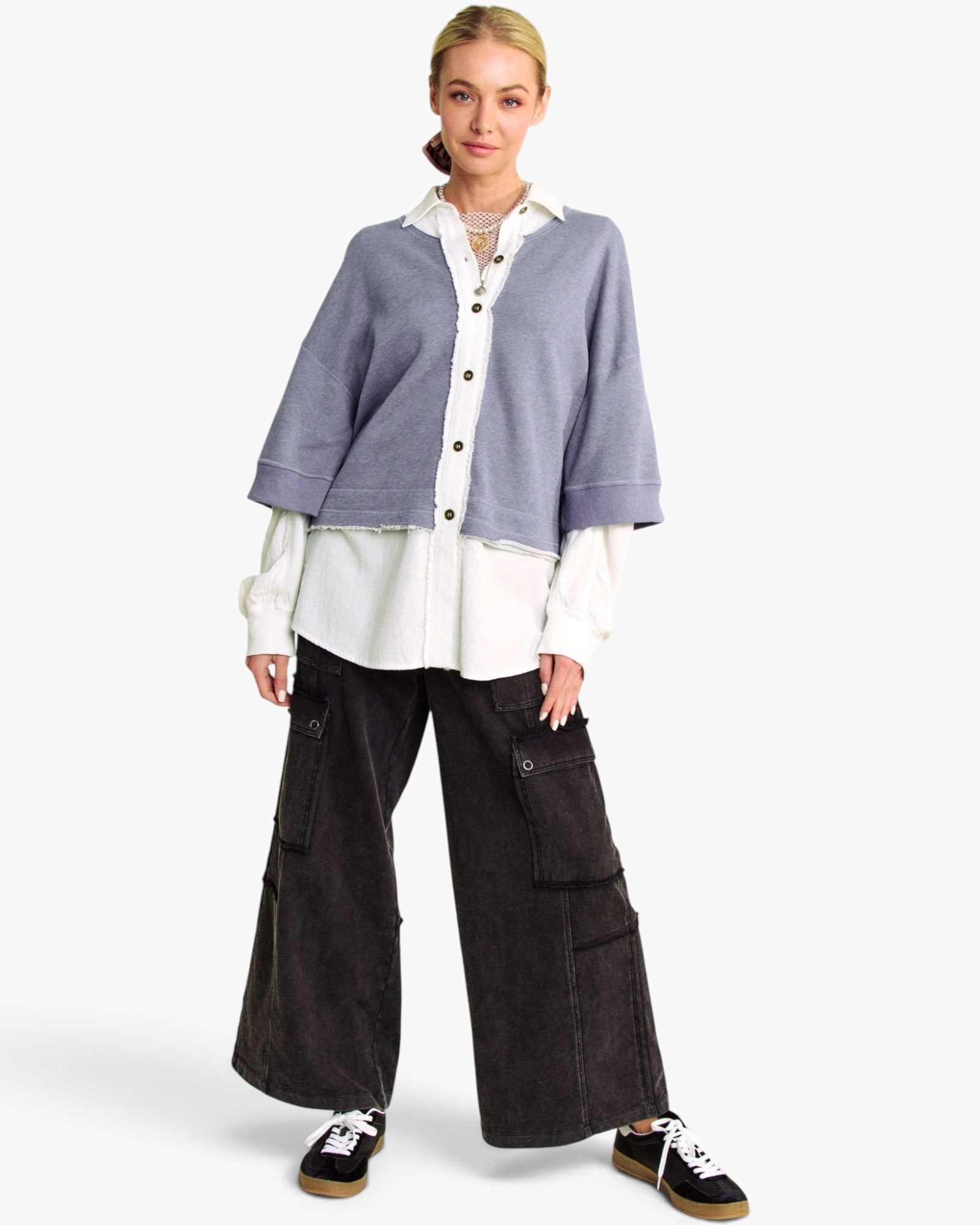 A model stands against a light gray studio background wearing a lavender cropped button-front cardigan over a white collared shirt, paired with wide-leg black cargo pants and black sneakers.