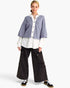 A model stands against a light gray studio background wearing a lavender cropped button-front cardigan over a white collared shirt, paired with wide-leg black cargo pants and black sneakers.