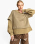 A woman modeling a loose-fit knit sweater with a solid taupe-brown body and ribbed knit sleeves and hem, paired with brown faux-leather pants against a plain white background.