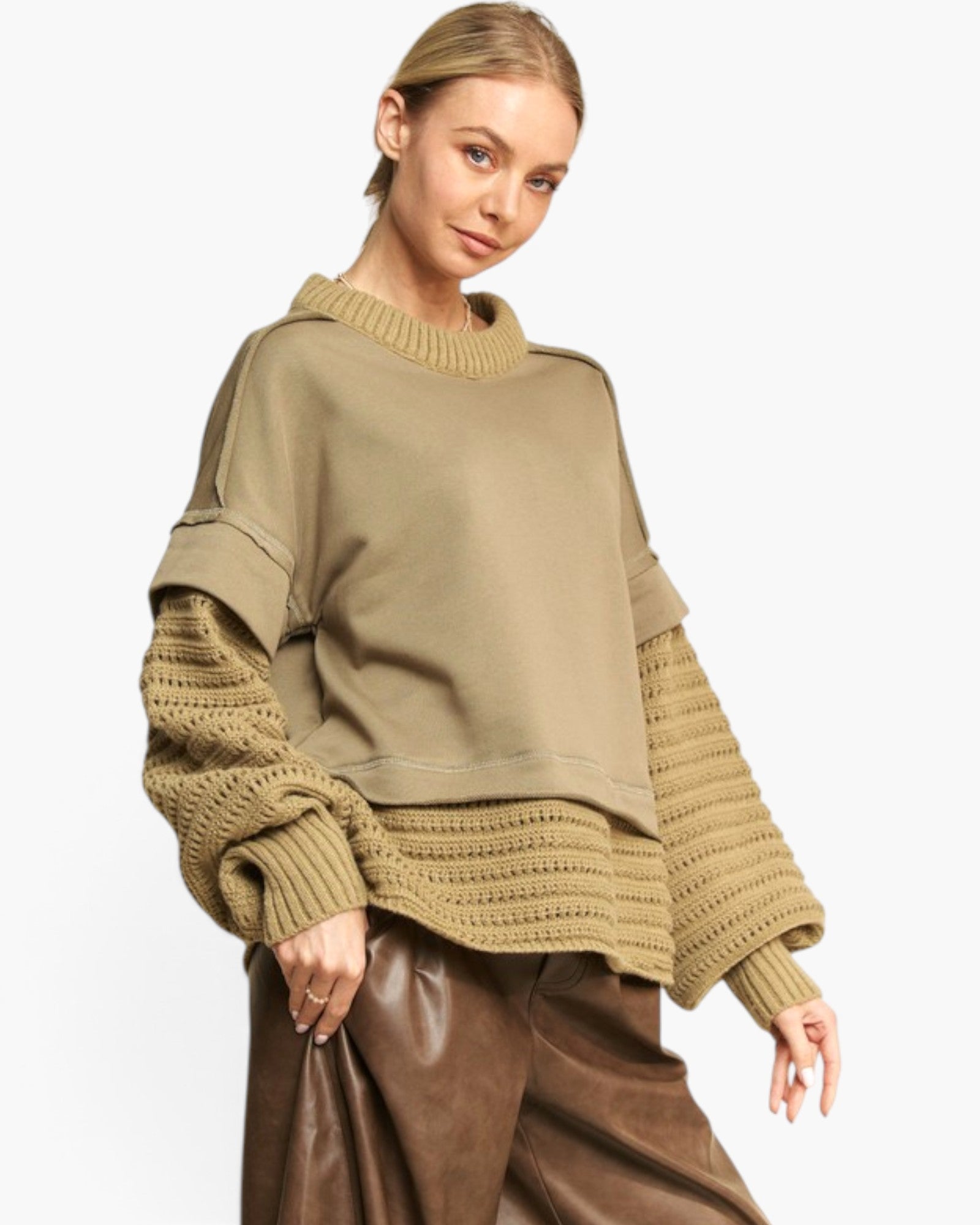 A woman wearing an oversized knit sweater with a solid taupe body and textured cable-knit sleeves and hem, paired with brown faux-leather pants, against a plain white studio background.