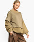 A woman wearing an oversized knit sweater with a solid taupe body and textured cable-knit sleeves and hem, paired with brown faux-leather pants, against a plain white studio background.
