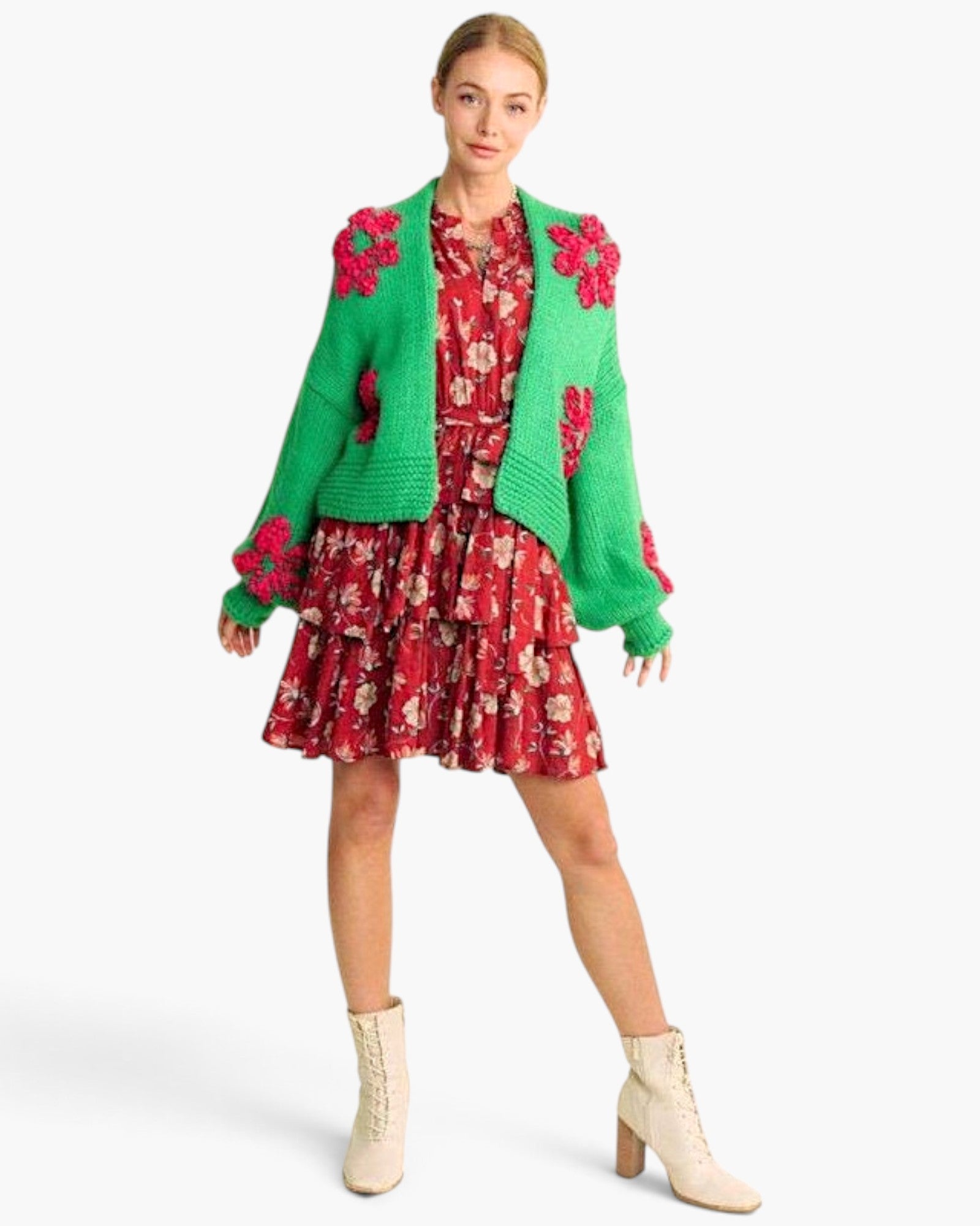 A model wearing a festive holiday-themed outfit featuring a bright green knit cardigan with red floral appliques, a red and pink floral mini dress, and cream lace-up ankle boots, set against a plain white studio background.
