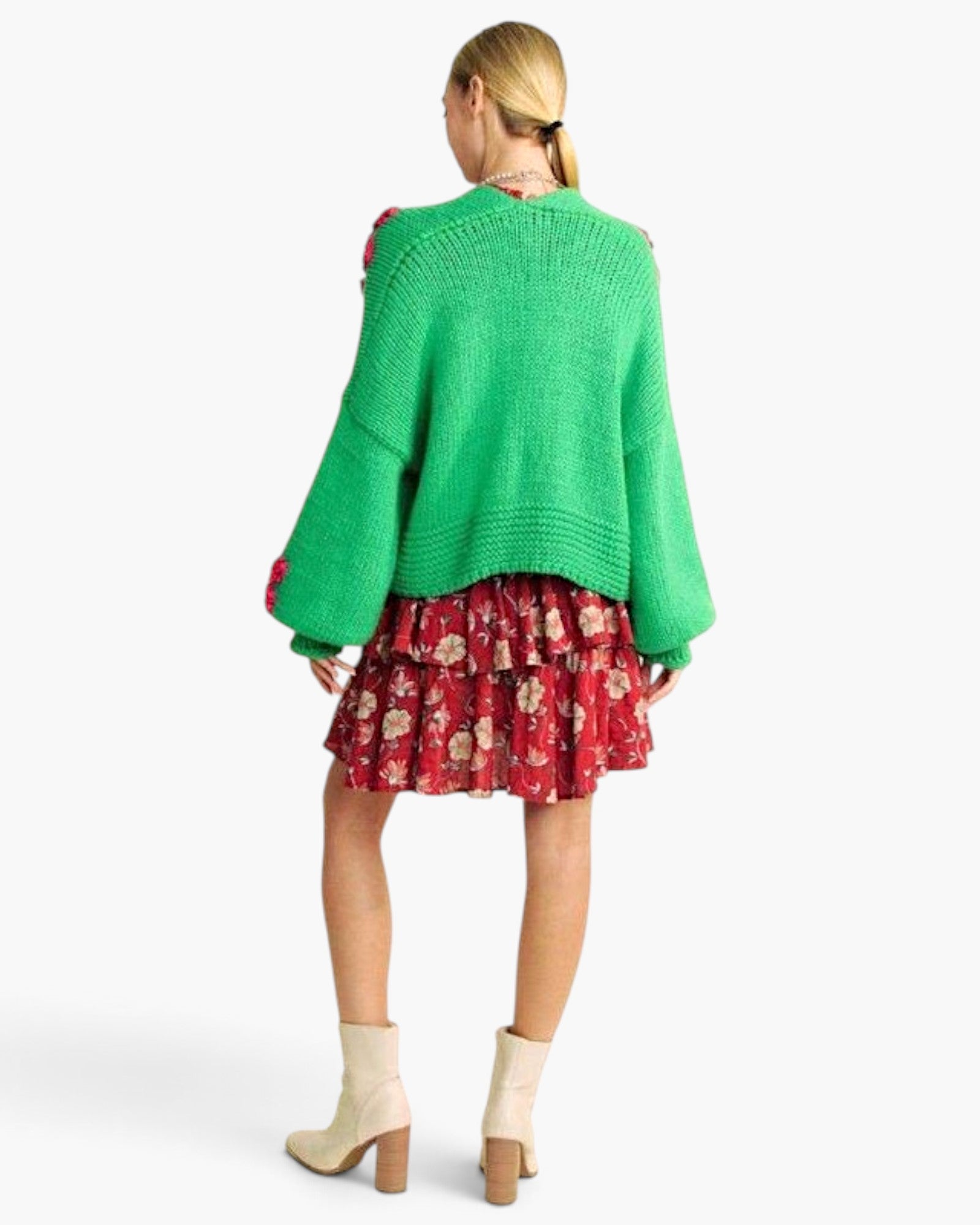 A woman shown from the back wearing a bright green knit sweater with red pom-pom accents, a red floral tiered skirt, and beige ankle boots against a light gray studio background.