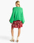A woman shown from the back wearing a bright green knit sweater with red pom-pom accents, a red floral tiered skirt, and beige ankle boots against a light gray studio background.