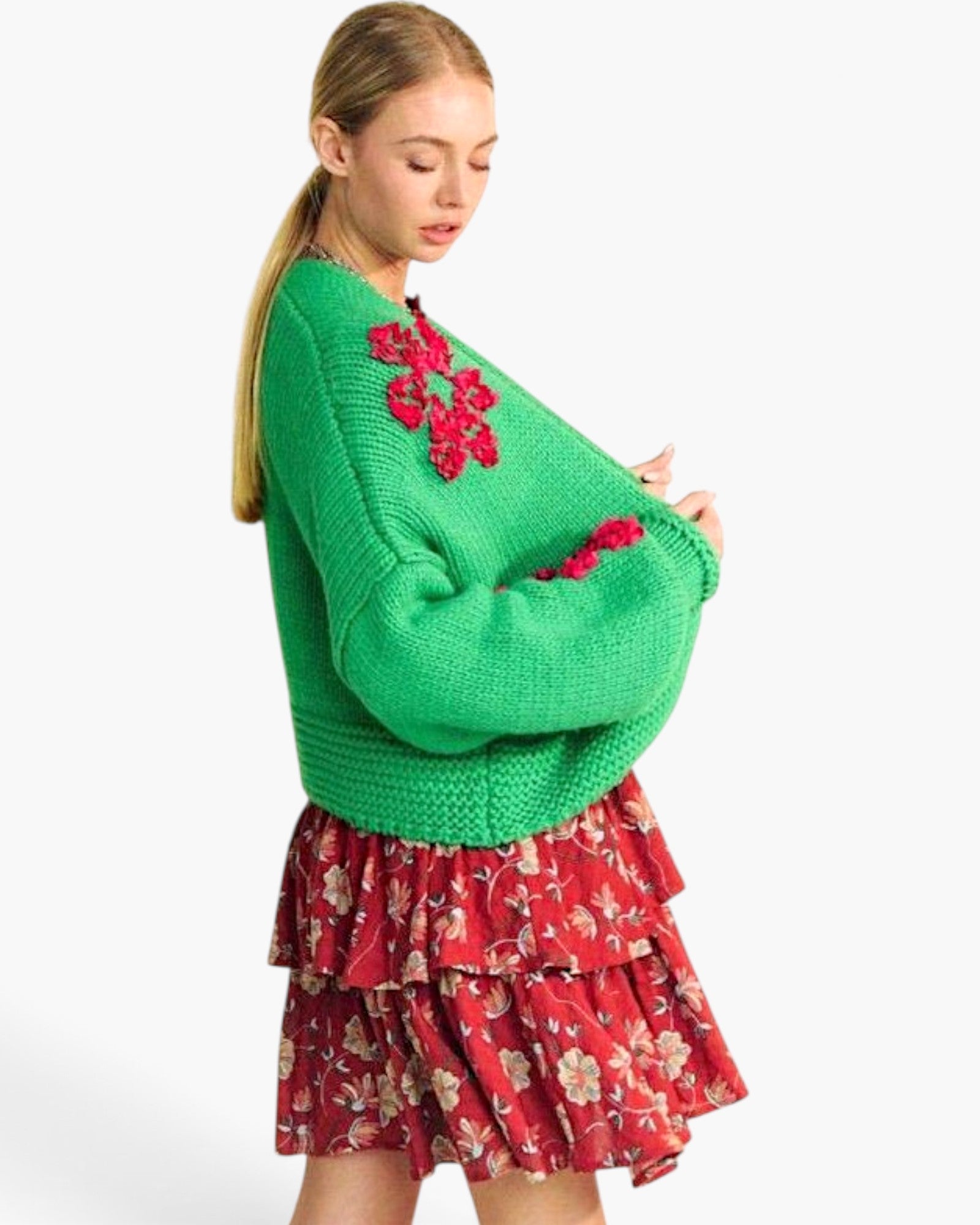 A woman modeling a bright green chunky knit sweater with red floral applique on the chest and sleeves, paired with a red floral tiered skirt, against a plain white background.