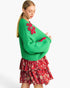 A woman modeling a bright green chunky knit sweater with red floral applique on the chest and sleeves, paired with a red floral tiered skirt, against a plain white background.
