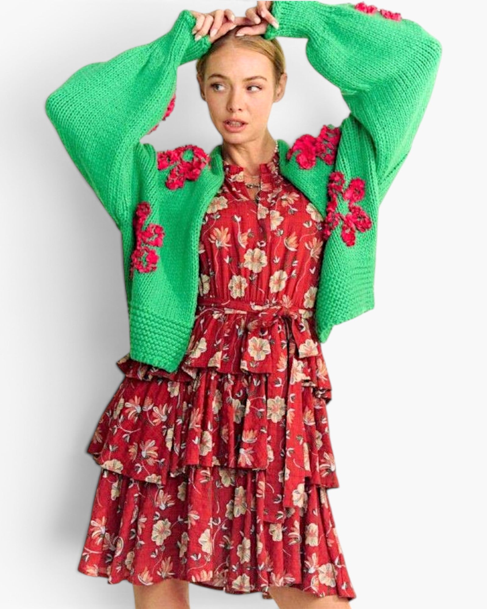 A woman modeling a bright green knit cardigan with pink floral embroidery over a red tiered floral dress, posed against a light gray studio background.