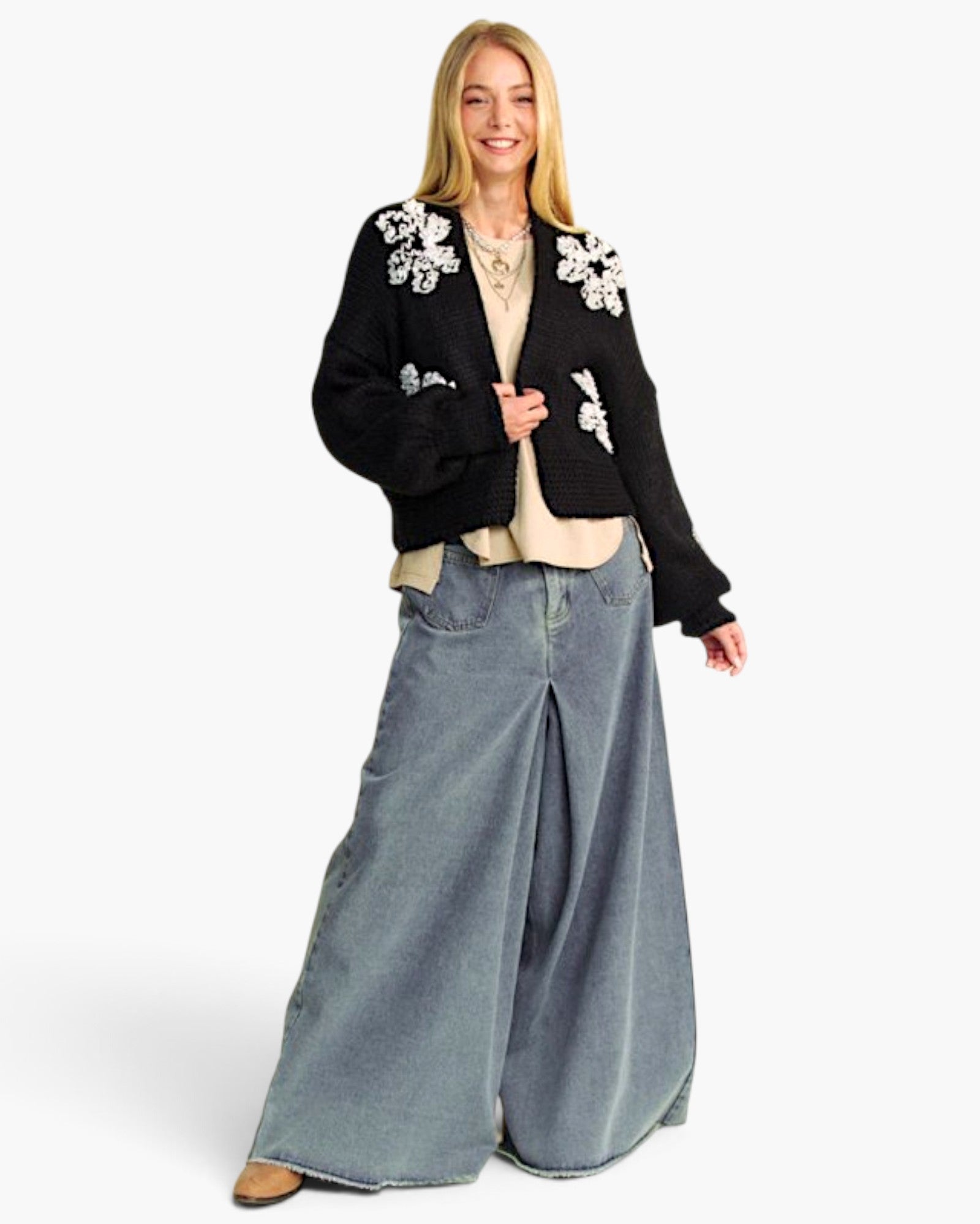 A woman modeling a black cropped knit cardigan with white floral embroidery on the shoulders, paired with wide-leg light blue jeans, a beige top, and brown shoes, against a plain white background.