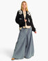 A woman modeling a black cropped knit cardigan with white floral embroidery on the shoulders, paired with wide-leg light blue jeans, a beige top, and brown shoes, against a plain white background.