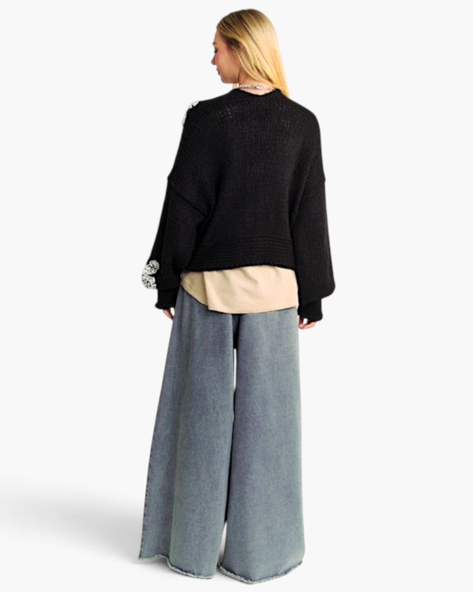 A woman shown from the back wearing a black cropped knit sweater, a beige shirt, wide-leg blue jeans, and a silver statement earring against a plain white background.