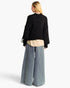 A woman shown from the back wearing a black cropped knit sweater, a beige shirt, wide-leg blue jeans, and a silver statement earring against a plain white background.