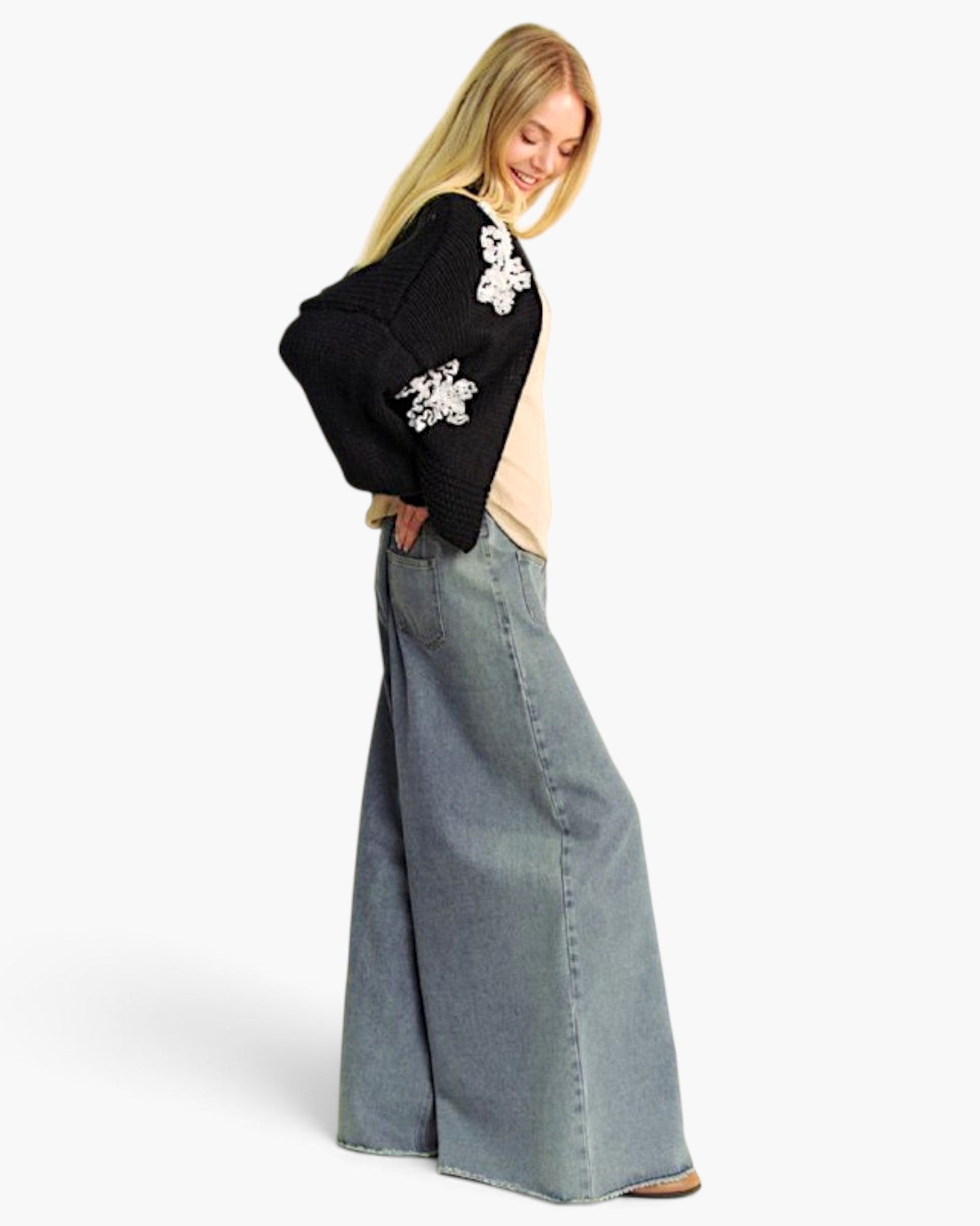 A woman modeling a black knit cardigan with white floral applique on the shoulders and sleeves, paired with a light beige top and wide-leg blue denim skirt, against a plain white studio background.
