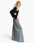 A woman modeling a black knit cardigan with white floral applique on the shoulders and sleeves, paired with a light beige top and wide-leg blue denim skirt, against a plain white studio background.