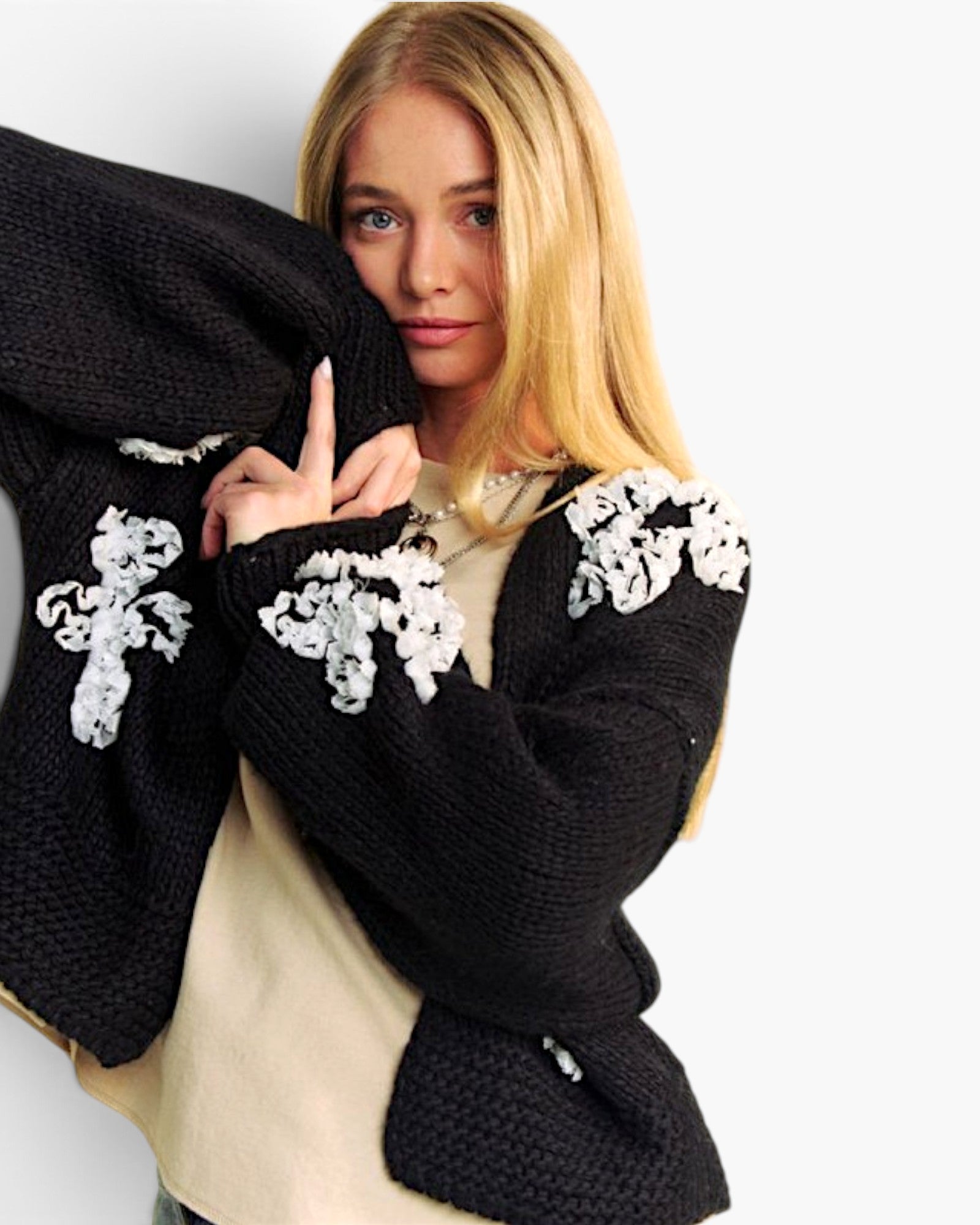 A blonde woman wearing a chunky black knit cardigan with white floral embroidery on the shoulders, layered over a beige top, against a plain white background.