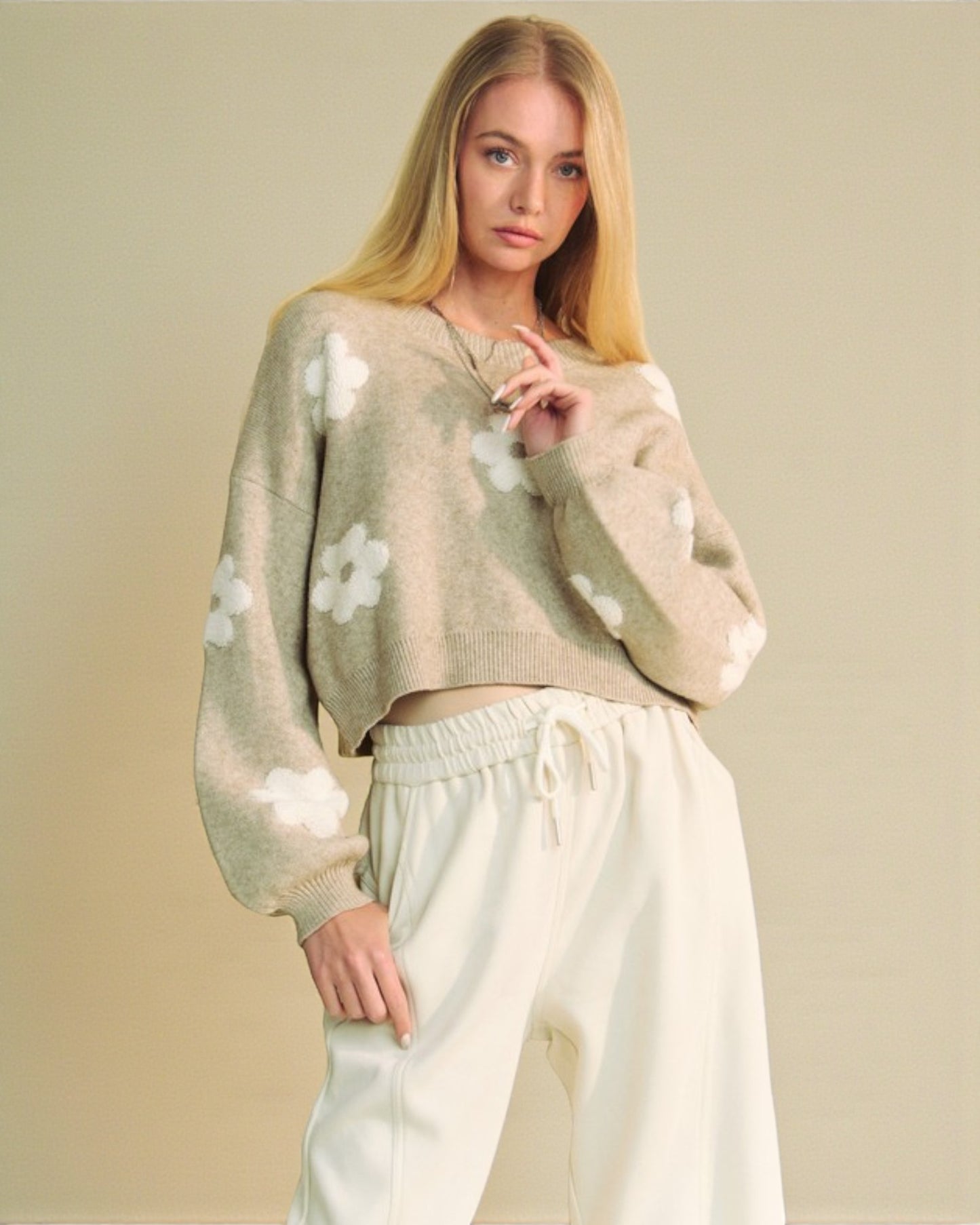 Woman wearing a beige sweater with white floral patterns and light-colored pants against a plain background