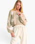 A blonde model wearing a cropped, long-sleeve knit sweater in a light beige color with white floral patterns, paired with cream-colored sweatpants, against a plain white studio background.