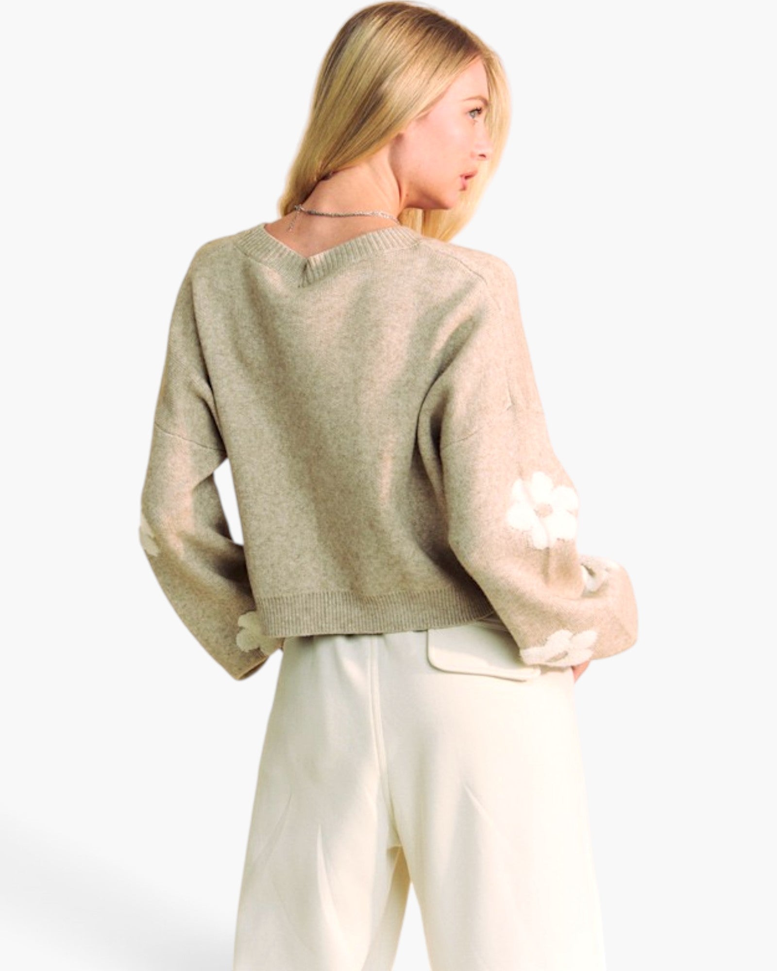 A woman shown from the back wearing a light beige knit sweater with white floral applique on the sleeves and white trousers, photographed against a clean white studio background.