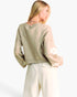 A woman shown from the back wearing a light beige knit sweater with white floral applique on the sleeves and white trousers, photographed against a clean white studio background.