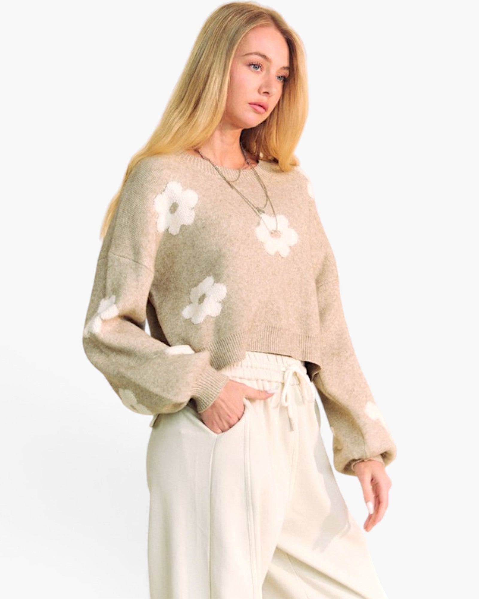 A woman modeling a beige knit sweater with large white flower motifs and cream drawstring pants against a plain white studio background.