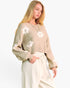 A woman modeling a beige knit sweater with large white flower motifs and cream drawstring pants against a plain white studio background.