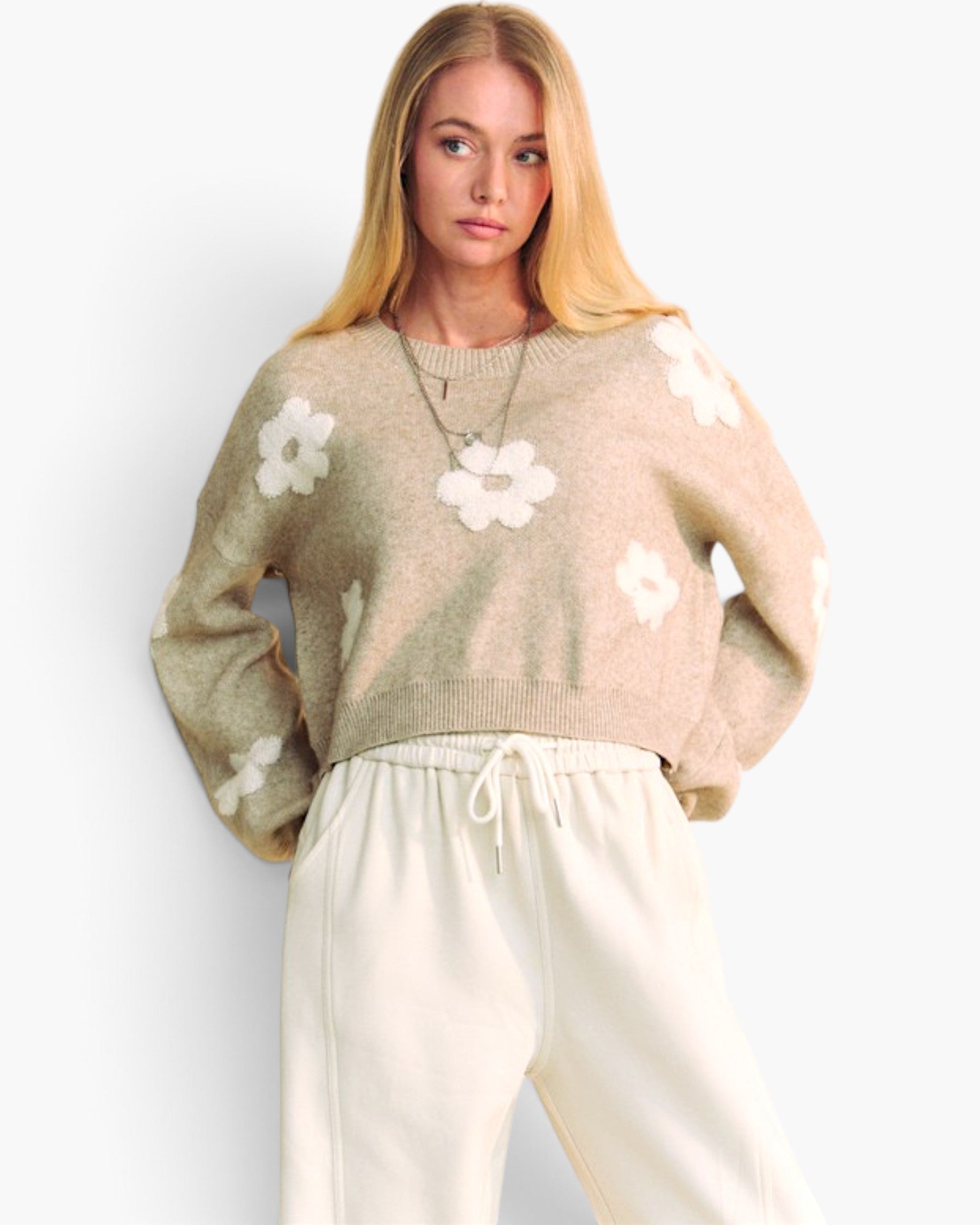 A woman modeling a cropped knit sweater in a light beige color with white floral motifs, paired with cream sweatpants, against a plain white background.