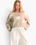 A woman modeling a cropped knit sweater in a light beige color with white floral motifs, paired with cream sweatpants, against a plain white background.