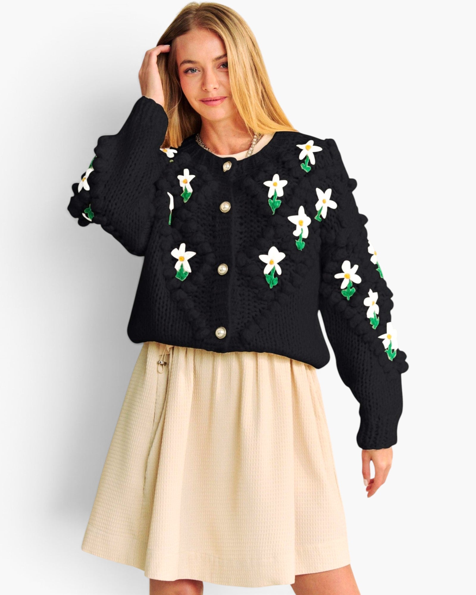 A woman modeling a black cable-knit cropped cardigan decorated with white daisy-like floral appliqués with green leaves, paired with a cream pleated skirt, against a light gray studio background.