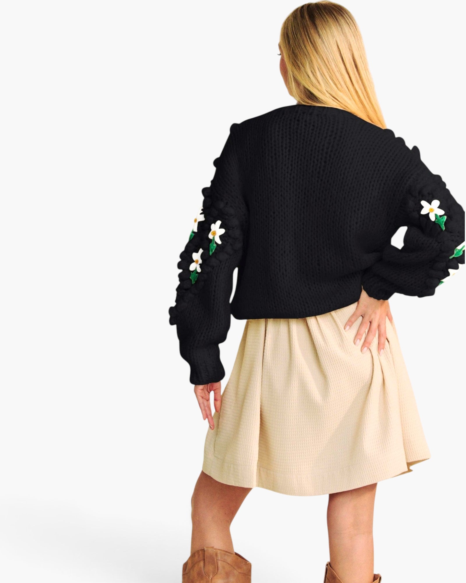A woman shown from the back wearing a black knit cropped sweater with ruffled sleeves and white floral applique, paired with a beige pleated skirt and brown boots, against a plain white studio background.