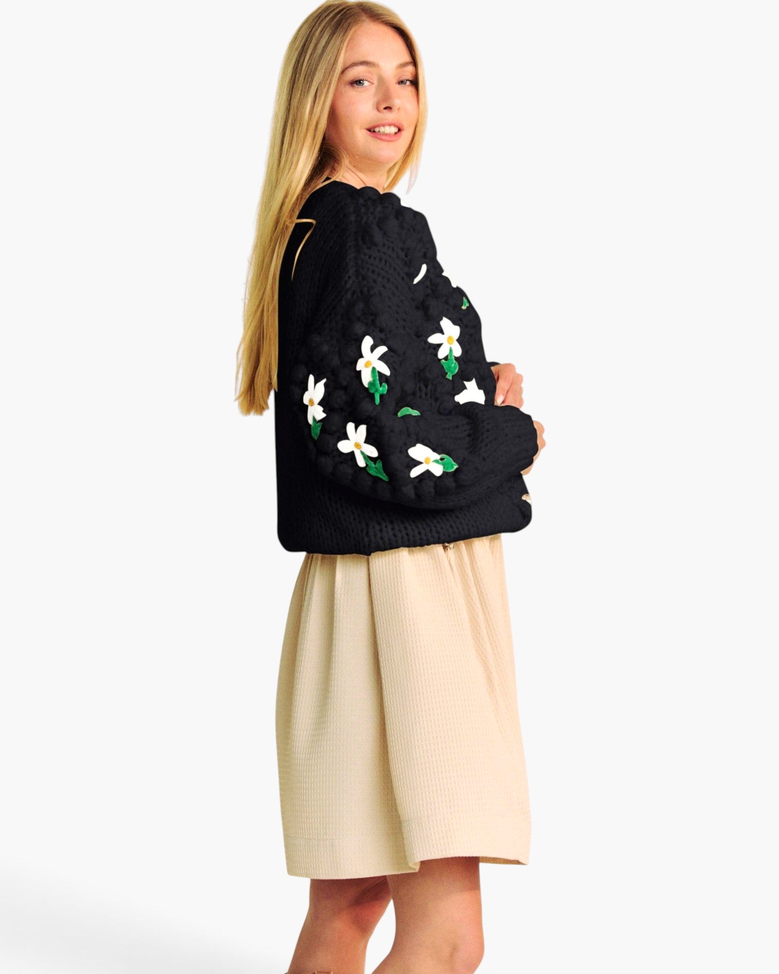 A young woman with long blonde hair is shown from a side-back angle, wearing a black knit sweater with white and green floral embroidery on the sleeves, paired with a beige skirt, against a plain white studio background.