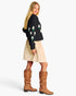 A woman modeling a black knit sweater with white and green floral embroidery on the sleeves, paired with a beige skirt and brown slouchy knee-high boots, against a plain white studio background.