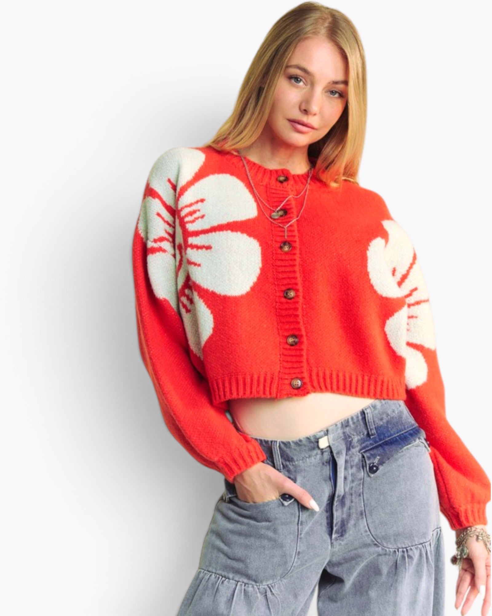 A woman wearing a cropped red knit cardigan with large cream floral motifs and gray jeans, standing against a plain light background.