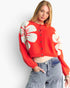 A woman wearing a cropped red knit cardigan with large cream floral motifs and gray jeans, standing against a plain light background.