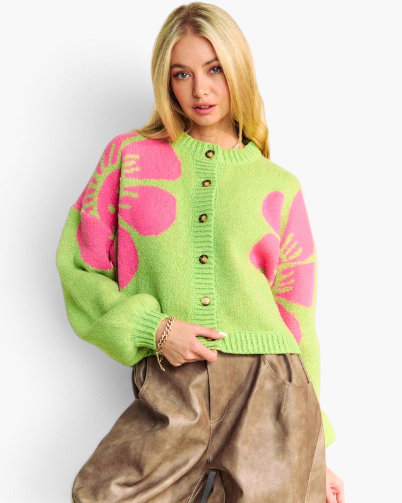 A blonde model wearing a bright lime green knit cardigan with large pink floral motifs and brown buttons, paired with brown faux-leather pants, against a plain white studio background.