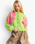 A blonde model wearing a bright lime green knit cardigan with large pink floral motifs and brown buttons, paired with brown faux-leather pants, against a plain white studio background.