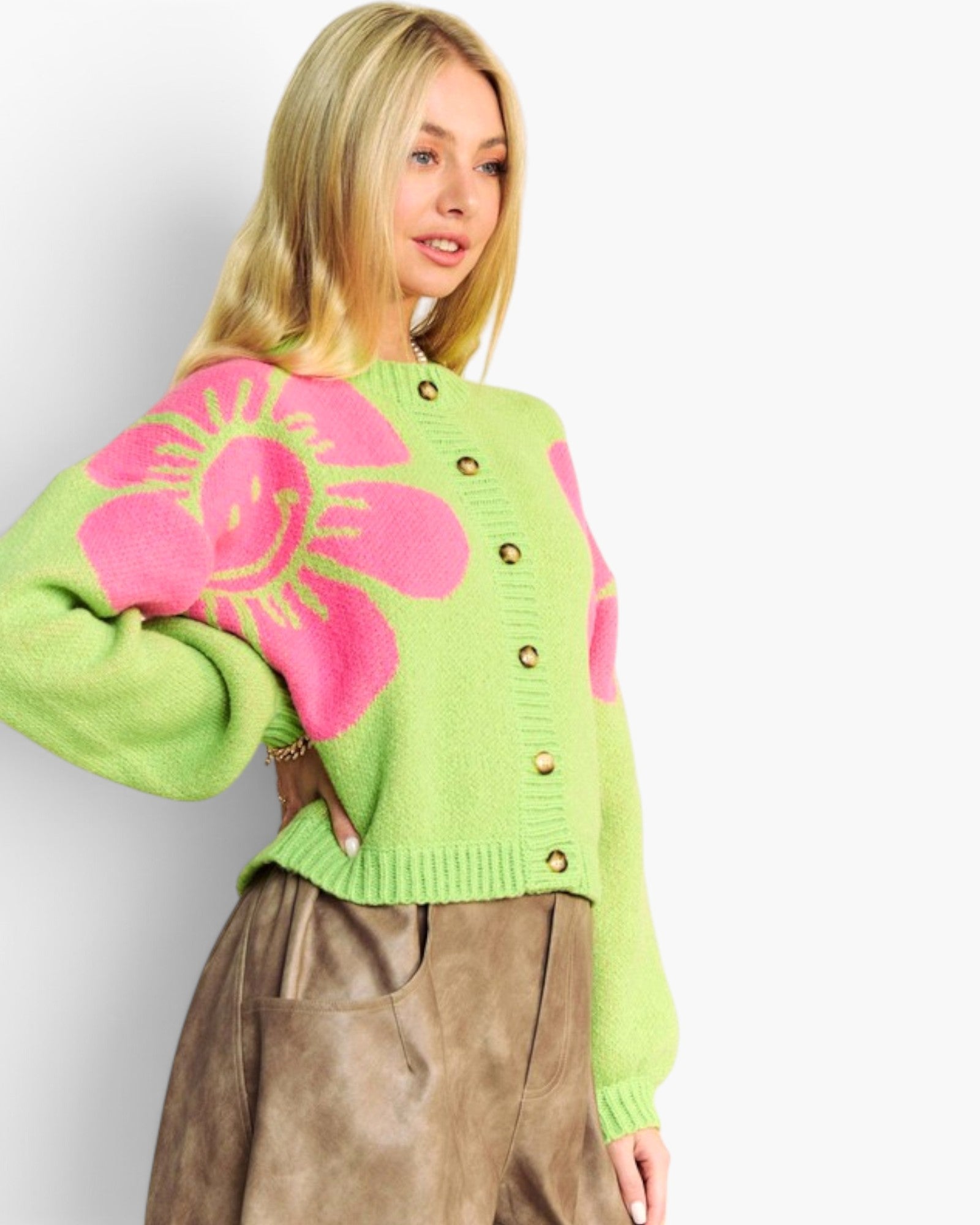 A woman wearing a bright lime green cardigan sweater with large pink sunflower motifs and gold buttons, paired with brown pants, against a plain light gray background.