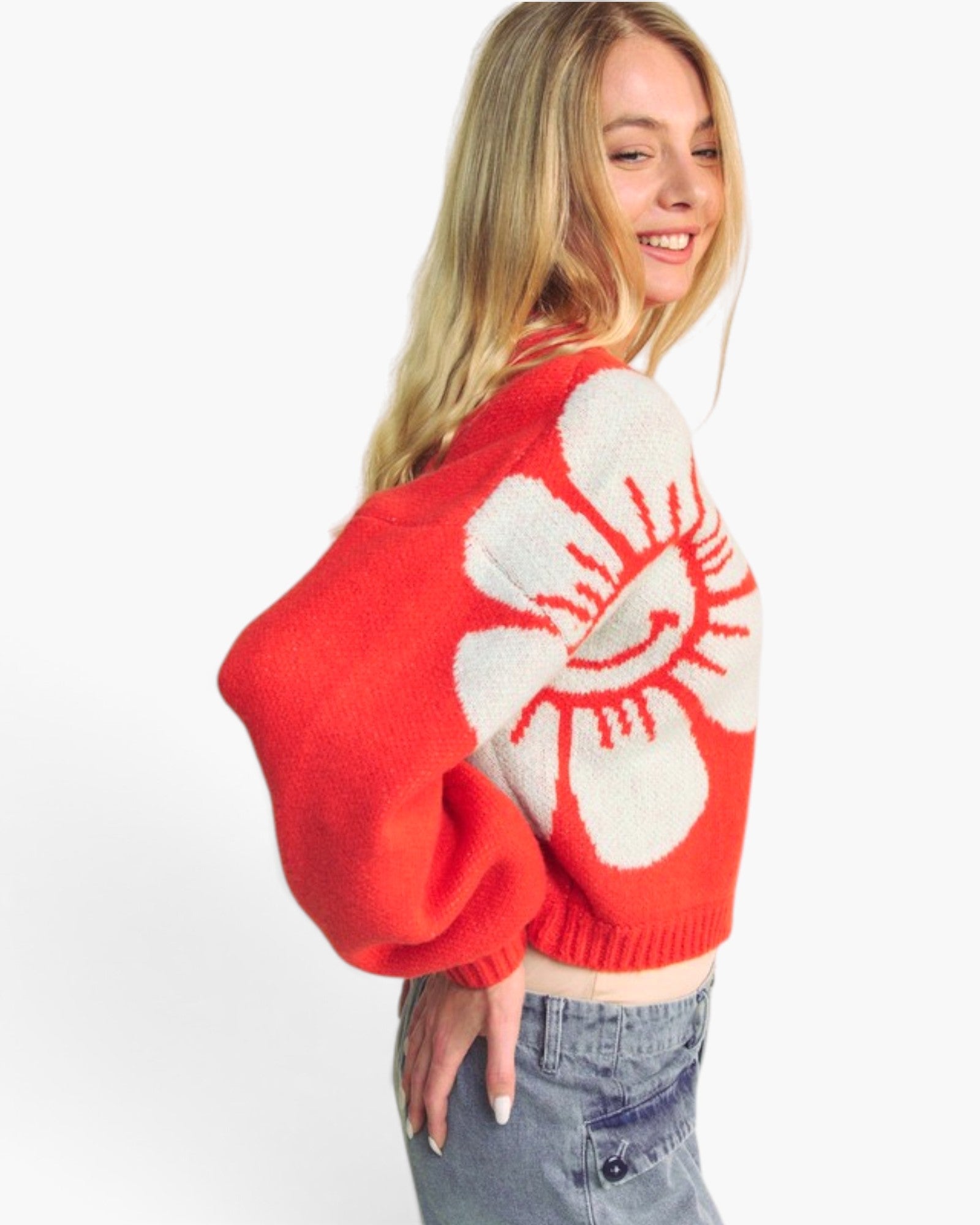 A woman wearing a bright red cropped knit sweater with a large white flower motif on the back, paired with light-wash denim jeans, photographed against a clean white studio background.