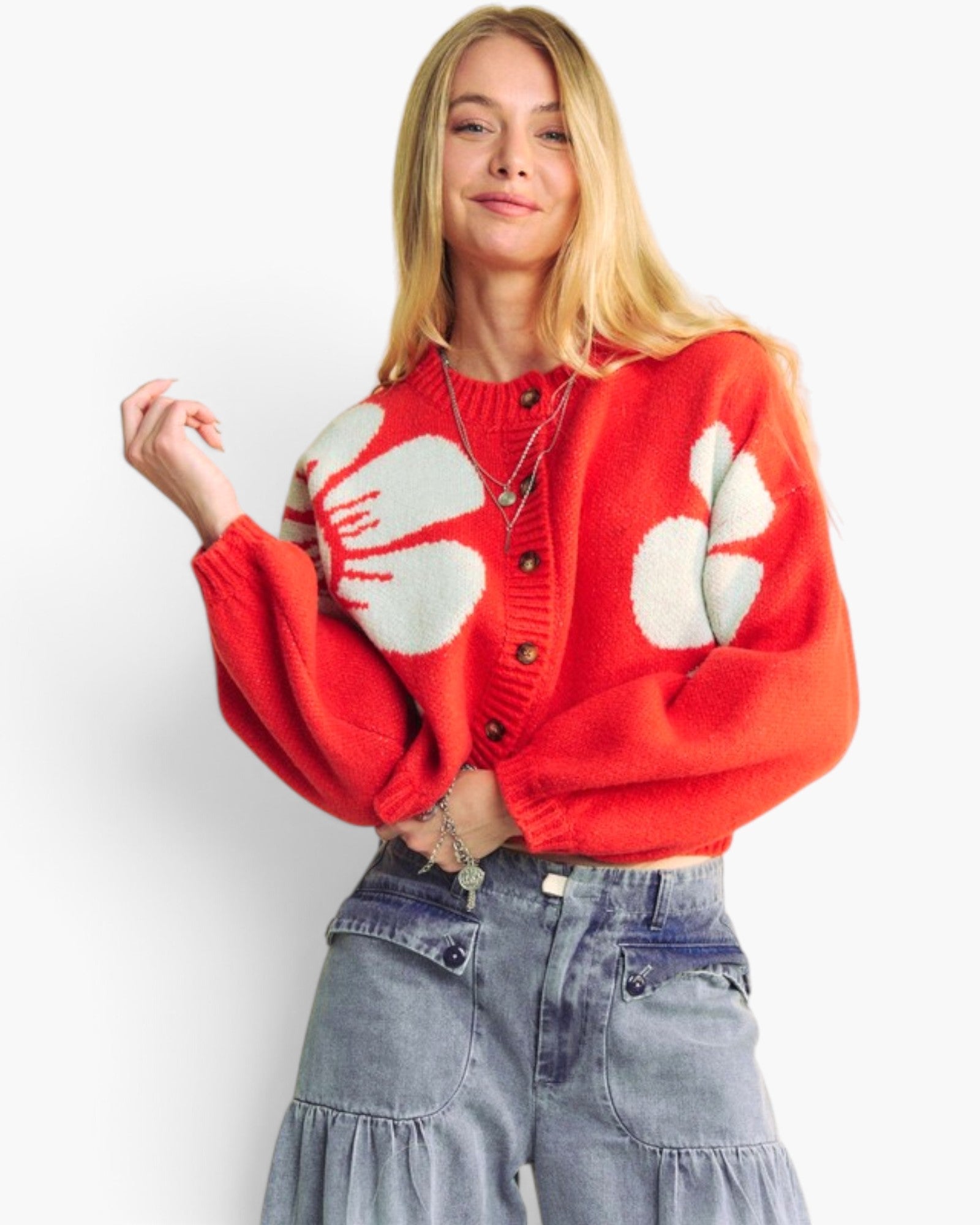A blonde woman wearing a bright red cropped cardigan with large white abstract floral motifs and light-wash denim jeans, photographed against a clean white studio background.