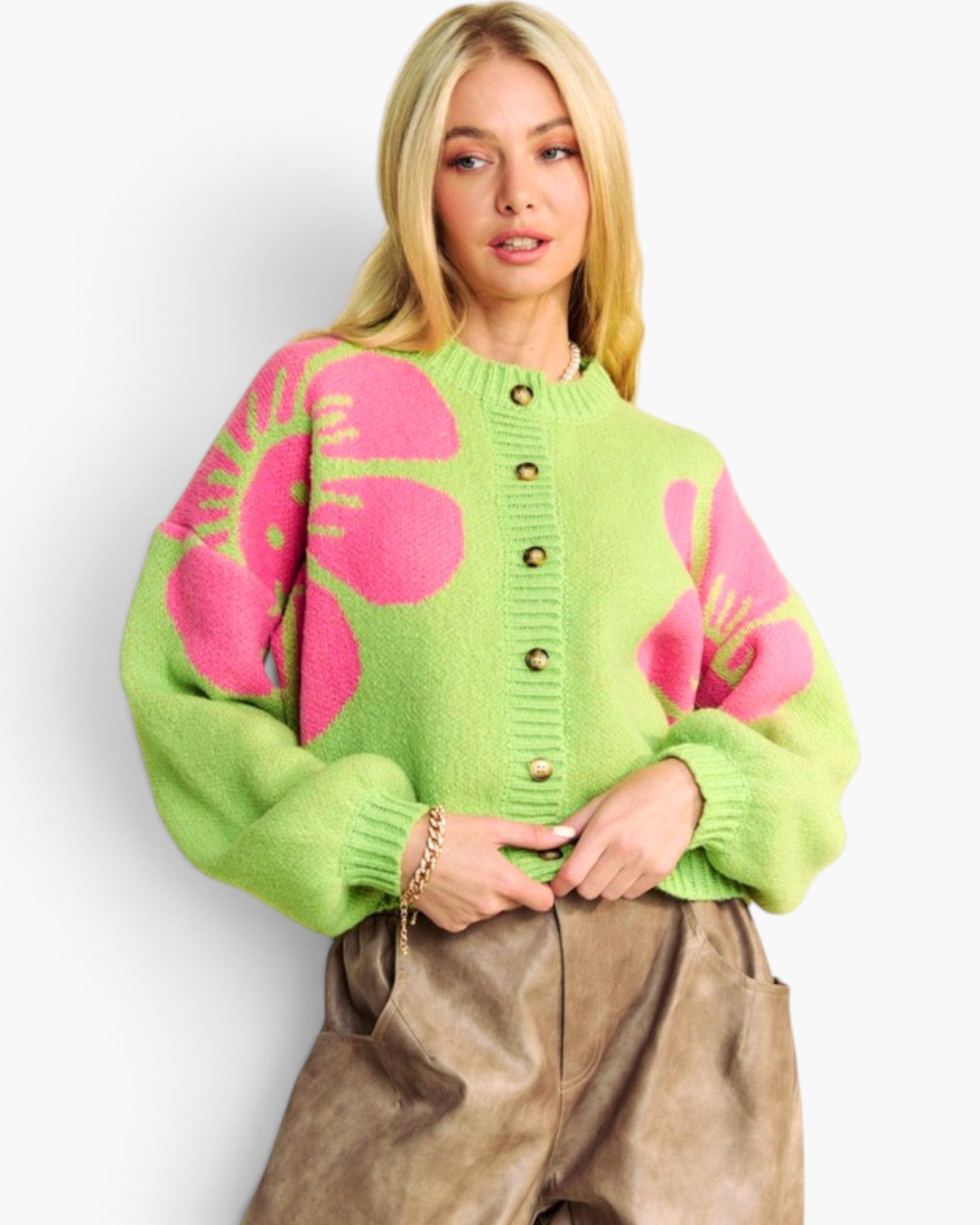 A blonde woman wearing a bright lime green knit cardigan with large pink floral motifs and brown buttons, paired with brown faux-leather pants, against a plain light gray background.