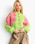 A blonde woman wearing a bright lime green knit cardigan with large pink floral motifs and brown buttons, paired with brown faux-leather pants, against a plain light gray background.