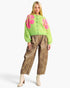A model wearing a bright lime green cardigan with pink floral patterns, brown wide-leg pants, and beige lace-up ankle boots, set against a plain light gray studio background.
