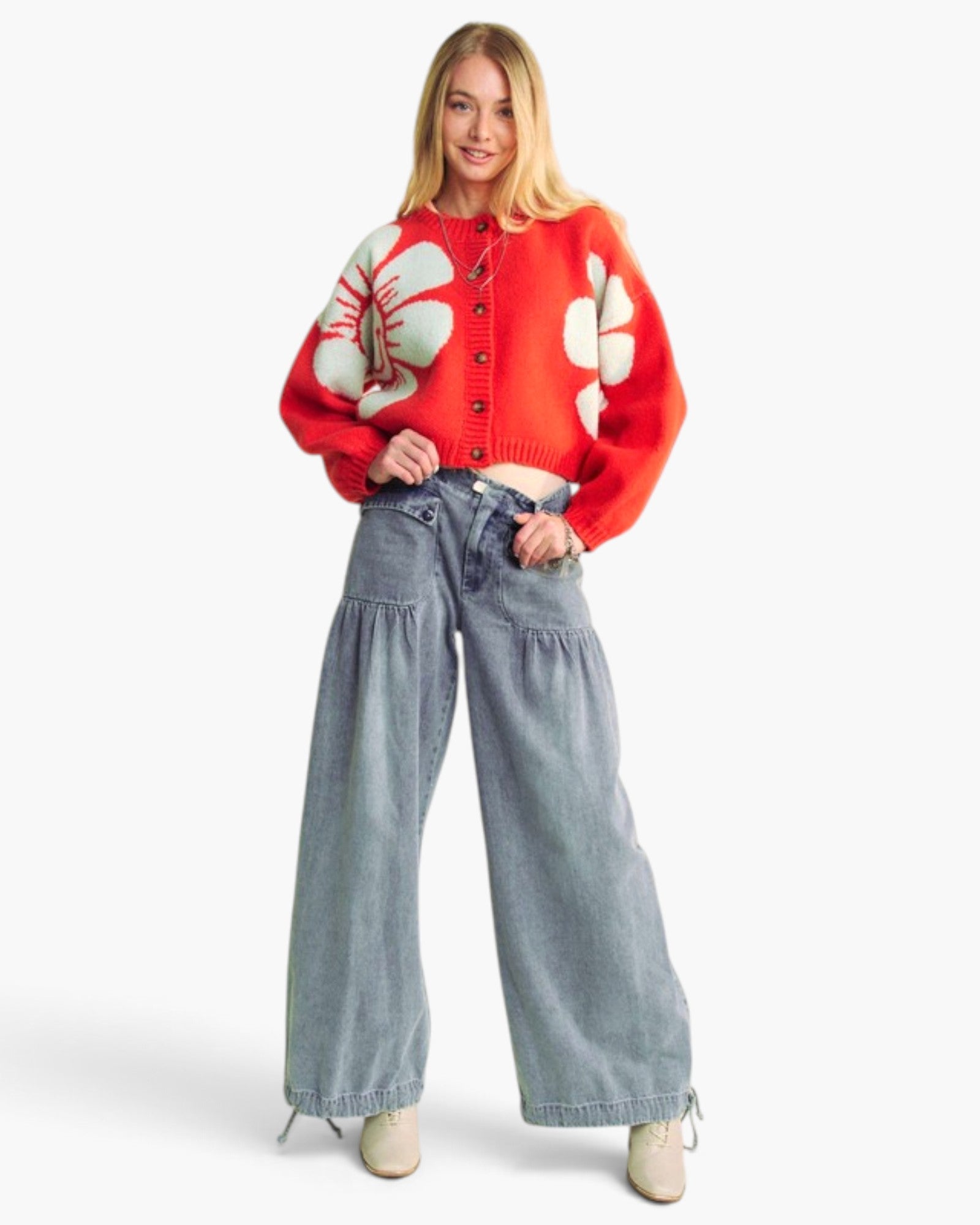 A woman modeling a vibrant red knit cardigan with large white floral motifs, paired with wide-leg light blue jeans and beige shoes, against a plain white studio background.