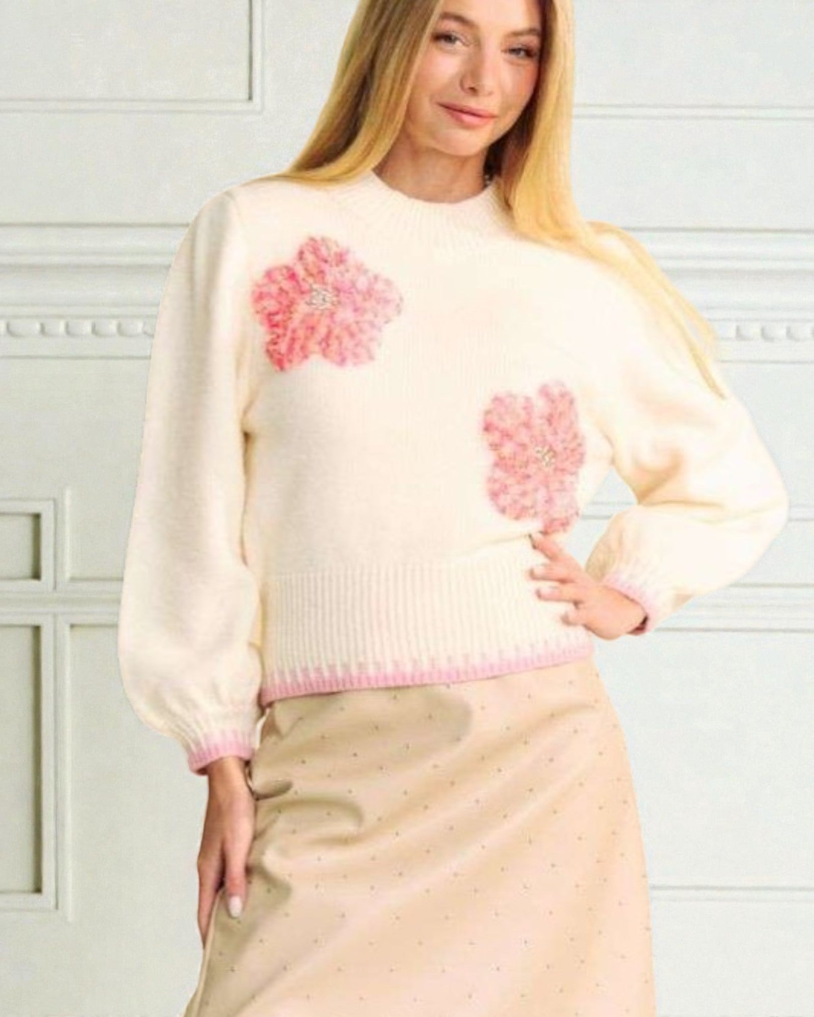 Woman wearing a cream sweater with pink floral patterns against a white wall.