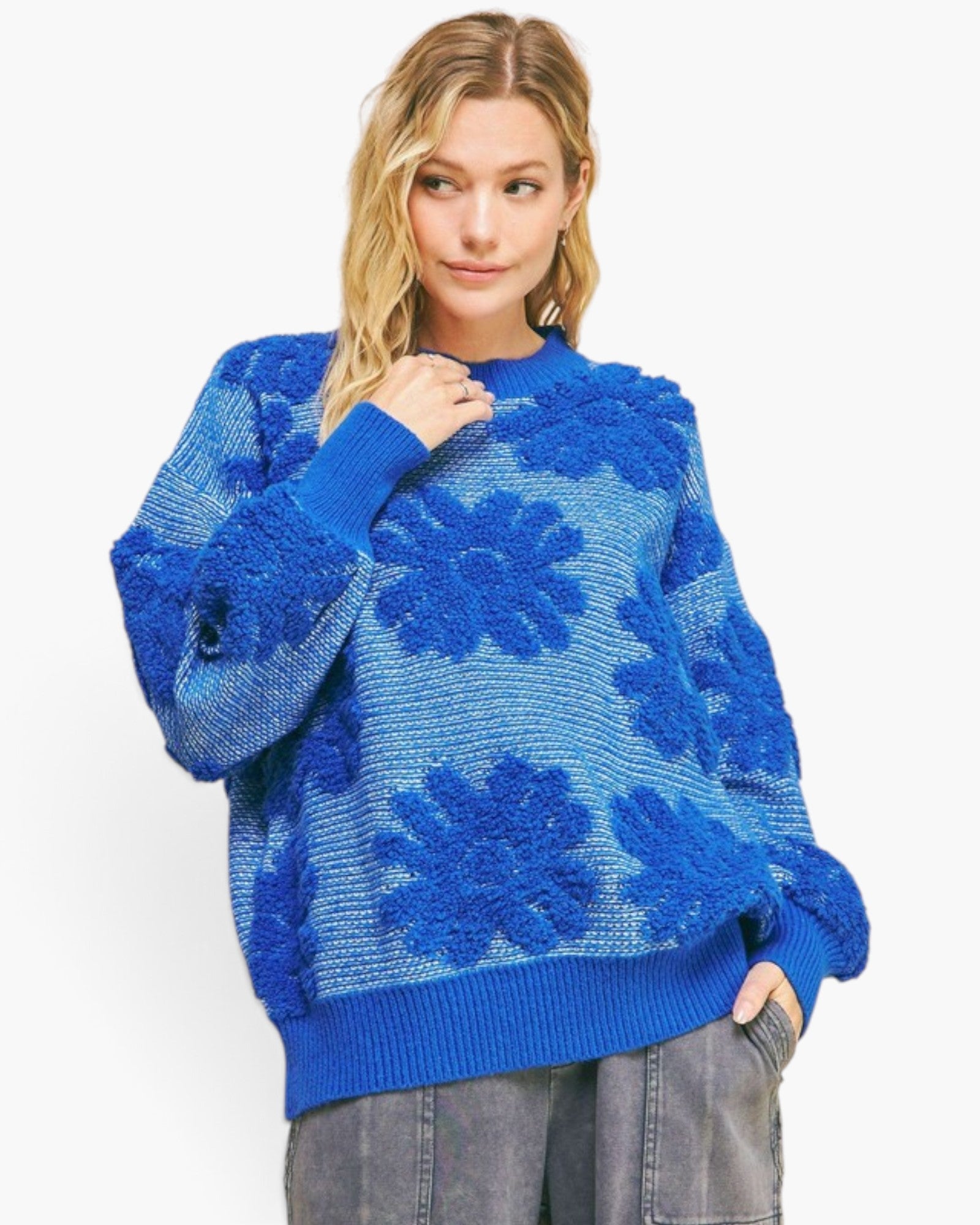 A woman wearing a vibrant blue textured knit sweater with raised floral patterns, paired with gray jeans, against a plain white background.