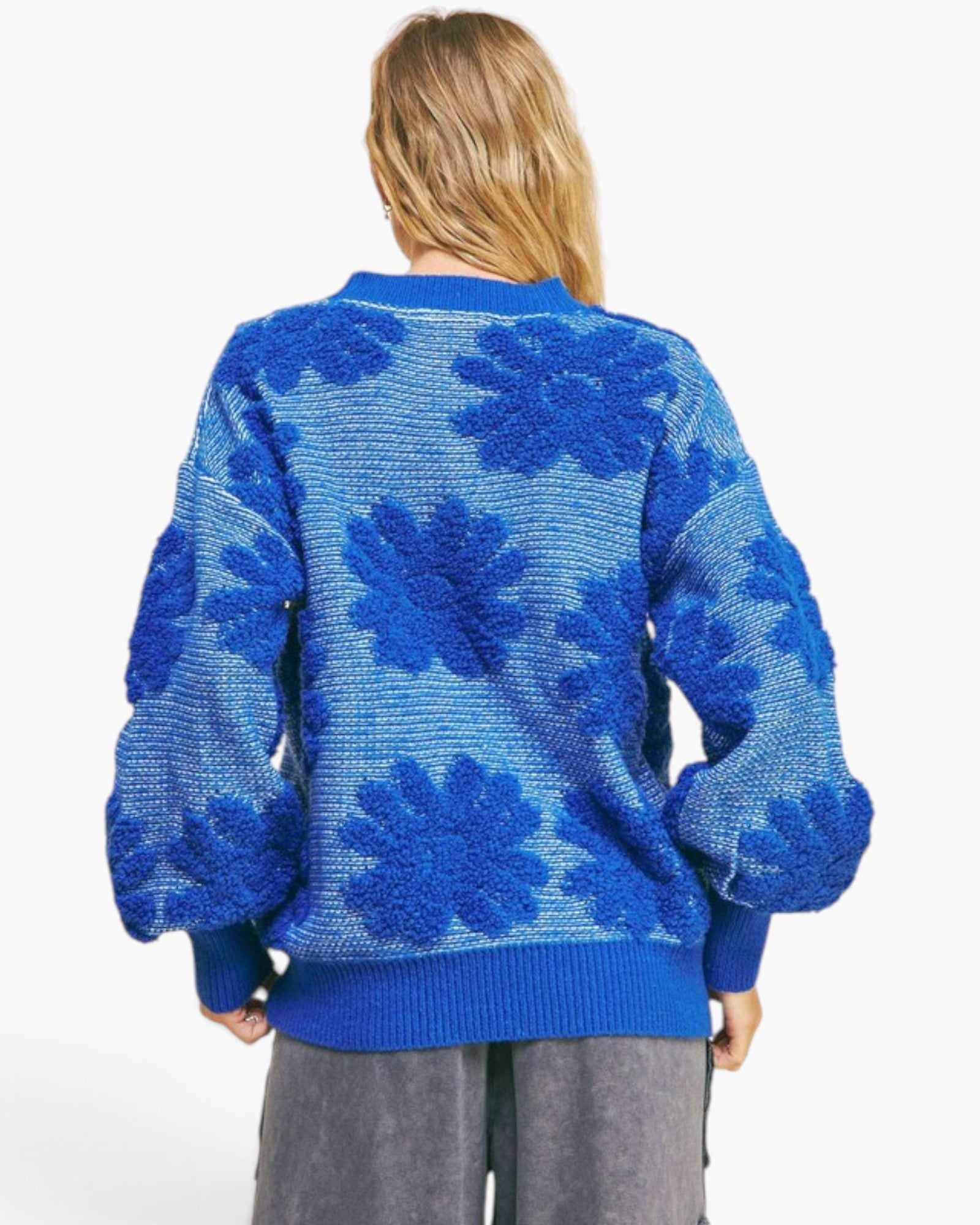 A woman viewed from behind wearing a blue textured knit sweater with large floral boucle patterns, paired with gray pants, against a plain white background.
