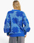 A woman viewed from behind wearing a blue textured knit sweater with large floral boucle patterns, paired with gray pants, against a plain white background.
