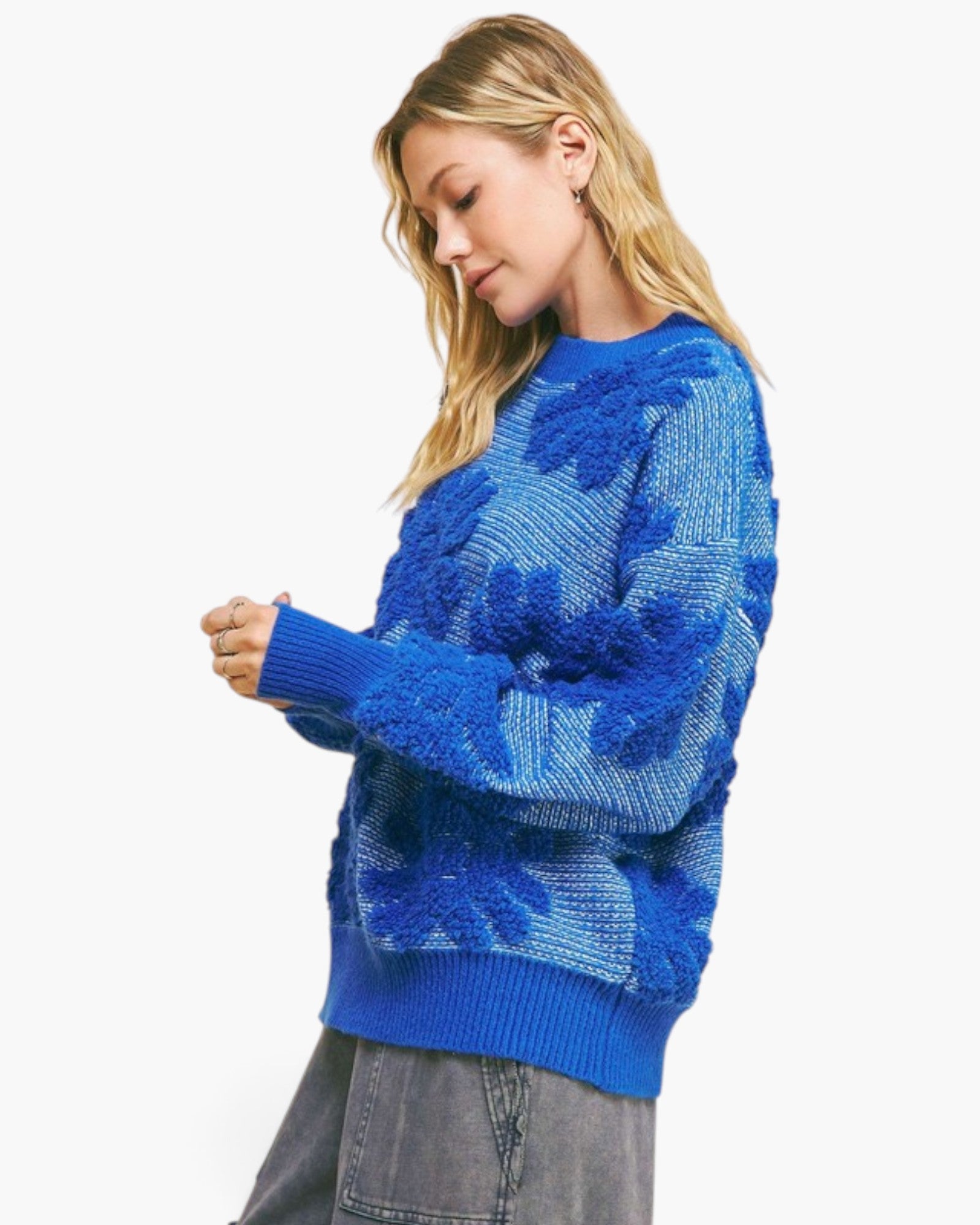 A woman wearing a textured royal blue knit sweater with raised floral patterns, paired with gray jeans, photographed against a clean white background.