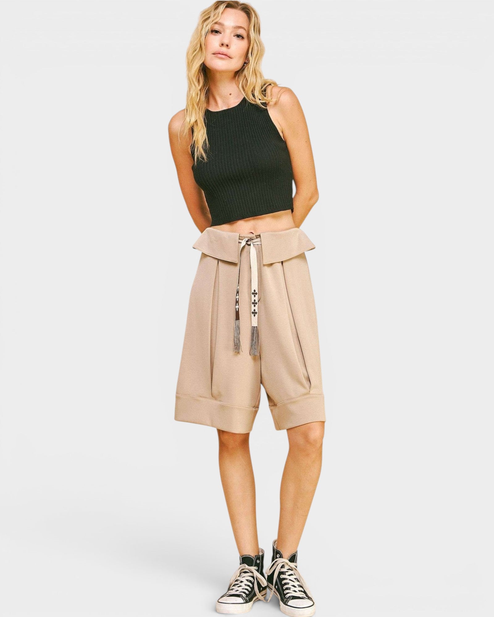 Woman wearing a black sleeveless top and beige high-waisted shorts with a belt on a white background