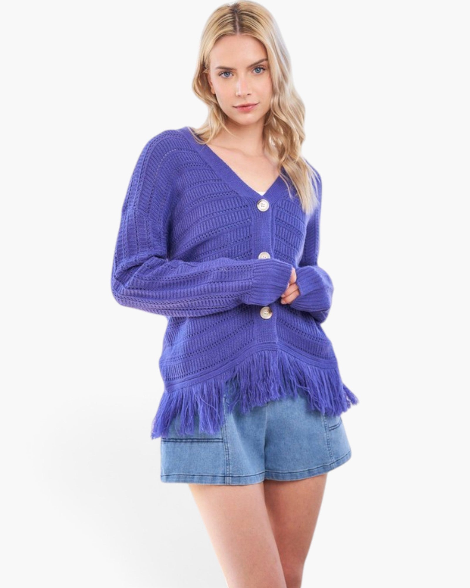 A woman modeling a purple chunky knit cardigan with a V-neck, button closure, and fringe hem, paired with denim shorts against a plain white background.